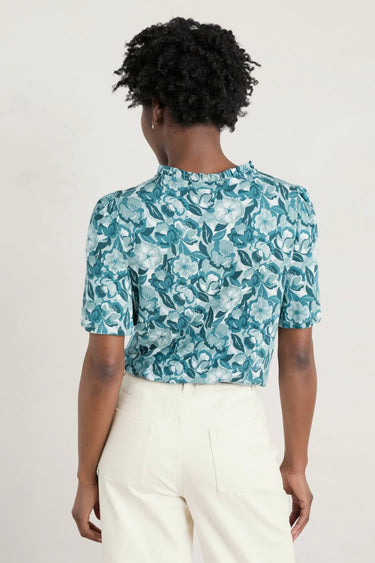 The back view of a woman wearing a teal floral Seasalt Anchor Hold Jersey Top and white pants.